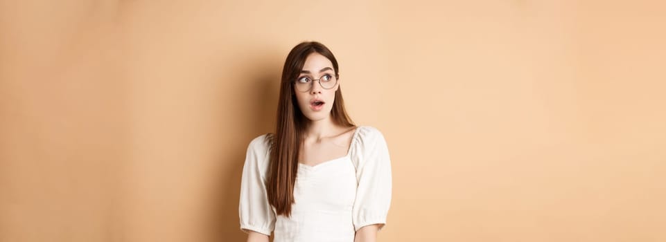 Excited young woman in glasses looking aside with opened mouth, gasping fascinated, standing on beige background.