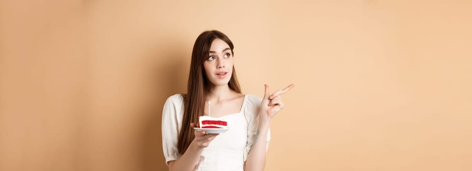 Thoughtful birthday girl holding cake with candle, thinking of wish and pointing left at logo, standing on beige background.