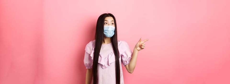 Coronavirus, quarantine and lifestyle concept. Excited asian teen girl in face mask pointing, looking left at logo with amazed face, checking out promotion, white background.