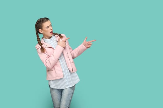 Portrait of shocked astonished teenager girl with braids wearing pink jacket pointing aside at advertisement area, space for promotion. Indoor studio shot isolated on green background.