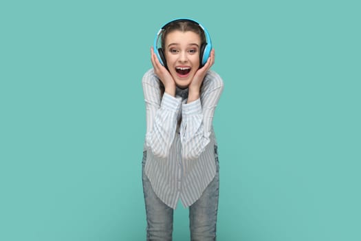 Portrait of amazed positive joyful teenager girl with braids listening music on headphones, looking at camera with happiness, hearing favorite song. Indoor studio shot isolated on green background.