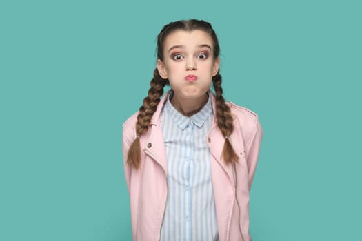 Portrait of funny teenager girl with braids wearing pink jacket blowing cheeks and puffing breath, has attractive look keeps silence with friends. Indoor studio shot isolated on green background.