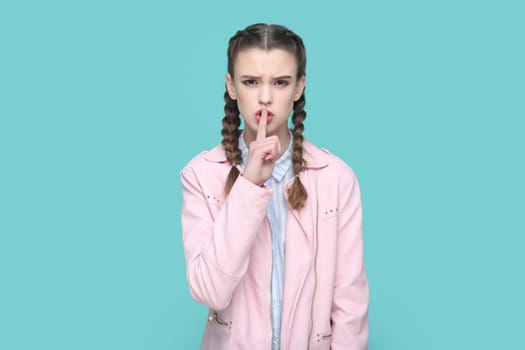 Portrait of serious teenager girl with braids wearing pink jacket keeps finger near lips, showing shh gesture, asking to keep silence. Indoor studio shot isolated on green background.