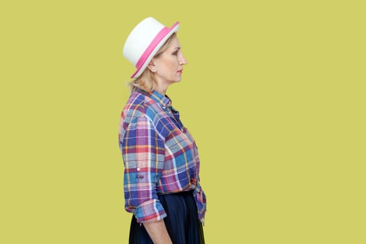 Side view portrait of serious strict mature woman wearing checkered shirt and hat standing looking ahead, being in bad mood. Indoor studio shot isolated on yellow background.