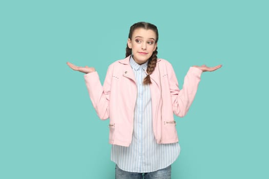Portrait of uncertain confused teenager girl with braids wearing pink jacket standing shrugging shoulders, spread hands, don't know answer. Indoor studio shot isolated on green background.