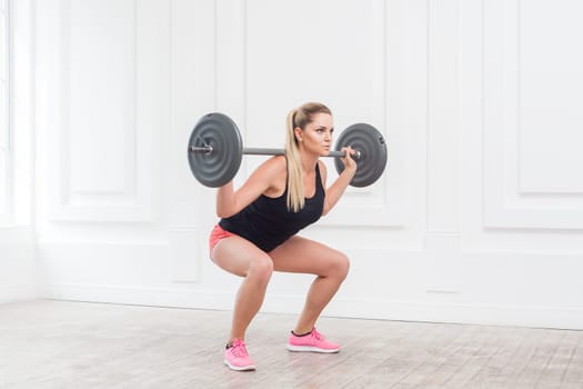 Side view portrait of sporty athletic beautiful woman in pink shorts and black top doing squats and exercising at the gym with the barbell on white wall background. Indoor studio shot.