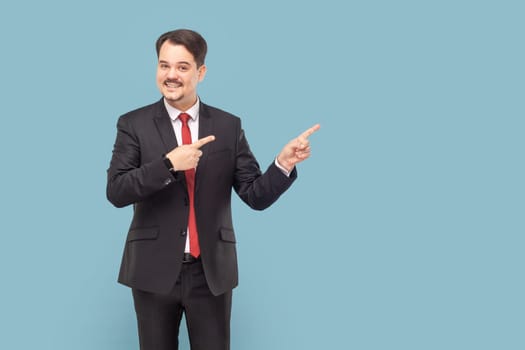 Smiling positive man with mustache standing pointing aside at empty space for advertisement or promotional text, wearing black suit with red tie. Indoor studio shot isolated on light blue background.