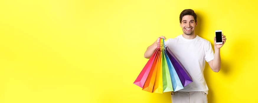 Young man holding shopping bags and showing mobile phone screen, money application, standing over yellow background.