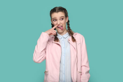 Portrait of funny childish teenager girl with braids wearing pink jacket standing with finger in nose, sticking tongue out, looking away. Indoor studio shot isolated on green background.