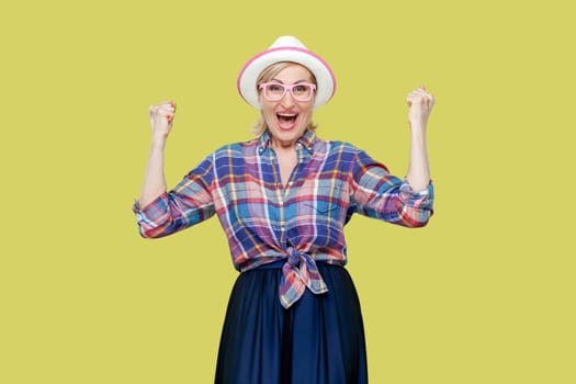 Overjoyed senior woman in checkered shirt, hat, eyeglasses clenches fists with happiness, opens mouth widely as shouts loudly, celebrates her success. Indoor studio shot isolated on yellow background.
