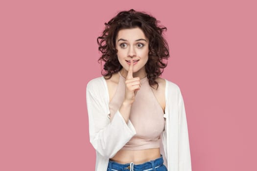 Portrait of positive smiling woman with curly hairstyle wearing casual style outfit showing shh gesture, asking to keep secret. Indoor studio shot isolated on pink background.