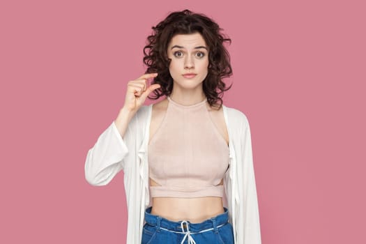 Portrait of confused disappointed beautiful woman with curly hairstyle wearing casual style outfit showing small size with fingers. Indoor studio shot isolated on pink background.