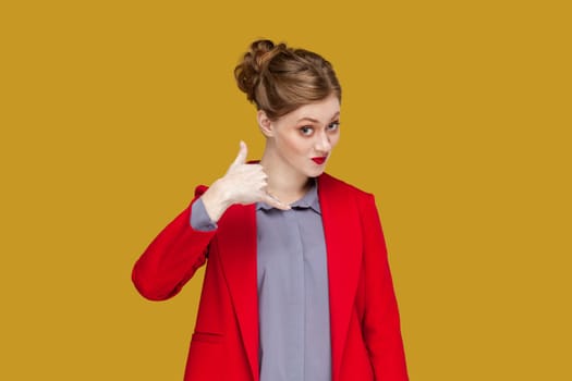 Portrait of flirting positive woman with red lips standing showing call me back gesture, looking at camera, wearing red jacket. Indoor studio shot isolated on yellow background.