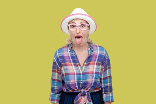 Portrait of childish mature woman wearing checkered shirt, hat and eyeglasses sticking out tongue, making funny grimace. Indoor studio shot isolated on yellow background.