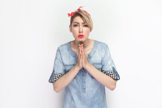 Portrait of sad hopeful blonde woman wearing blue denim shirt and red headband standing with praying gesture, asking to forgive her. Indoor studio shot isolated on gray background.