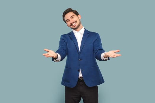 Portrait of friendly positive man with mustache standing with spread hands, saying welcome, take for free, wearing white shirt and jacket. Indoor studio shot isolated on light blue background.