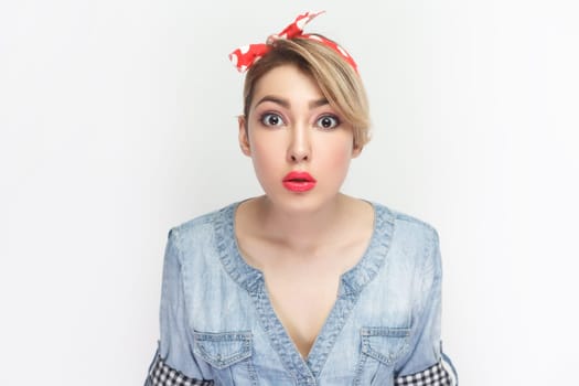 Portrait of astonished surprised blonde woman wearing blue denim shirt and red headband standing looking at camera with big eyes, being amazed. Indoor studio shot isolated on gray background.