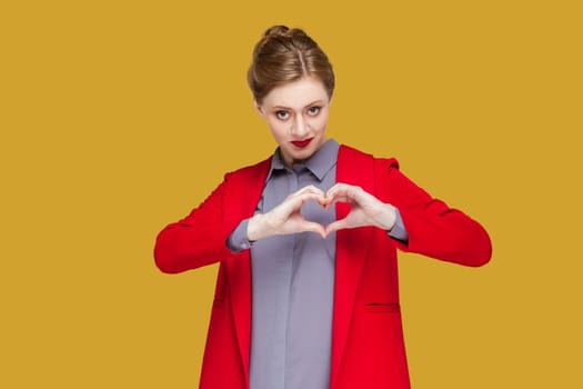 Portrait of romantic flirting woman with red lips standing showing heart shape with hands, looking at camera, expressing devotion, wearing red jacket. Indoor studio shot isolated on yellow background.