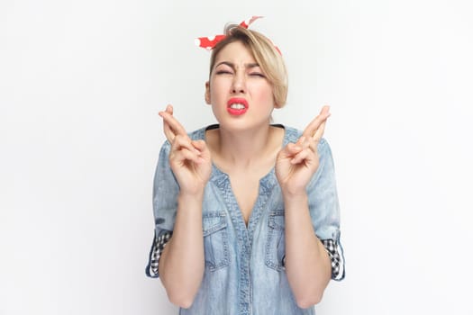 Portrait of hopeful attractive blonde woman wearing blue denim shirt and red headband standing with crossed fingers, praying for good luck. Indoor studio shot isolated on gray background.