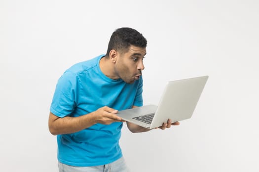 Portrait of shocked surprised unshaven man wearing blue T- shirt standing sees something astonished at laptop screen, looking at monitor with big eyes. Indoor studio shot isolated on gray background.