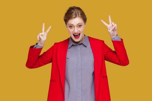 Portrait of excited extremely happy woman with red lips standing showing v sign with both hands, celebrating her success, wearing red jacket. Indoor studio shot isolated on yellow background.