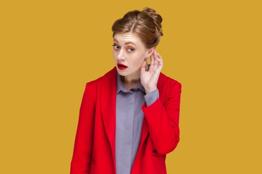 Portrait of curious woman with red lips standing with hand near ear listening something interesting, private conversation, wearing red jacket. Indoor studio shot isolated on yellow background.