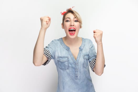 Portrait of extremely happy cheerful blonde woman wearing blue denim shirt and red headband standing clenched fists, celebrating victory. Indoor studio shot isolated on gray background.