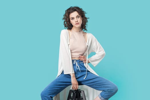 Portrait of winsome young adult woman with wavy hair sitting on chair, looking at camera confident expression, wearing stylish outfit. Indoor studio shot isolated on blue background.