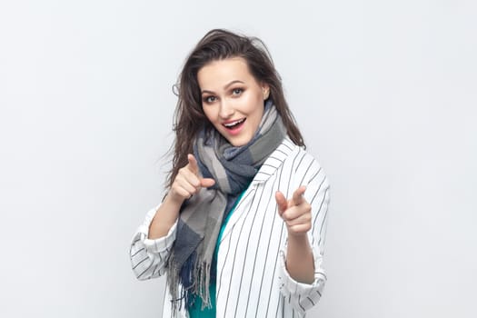 Portrait of overjoyed happy positive brunette woman pointing at camera with index fingers, choosing you, wearing striped jacket and scarf. Indoor studio shot isolated on gray background.