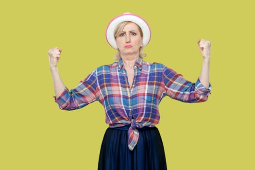 Portrait of mature woman wearing checkered shirt and hat raises hands and shows muscles, feels proud about her achievements in gym, smiles broadly. Indoor studio shot isolated on yellow background.