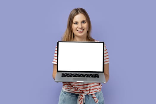 Portrait of happy joyful blond woman wearing striped T-shirt holding laptop with white empty display with empty space for advertisement. Indoor studio shot isolated on purple background.