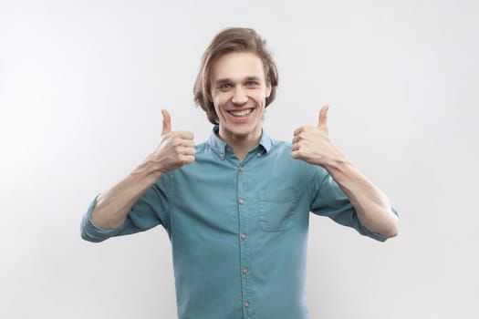 Smiling satisfied joyful attractive young man standing showing like gesture, thumbs up, recommend something, positive feedback, wearing blue shirt. Indoor studio shot isolated on gray background.