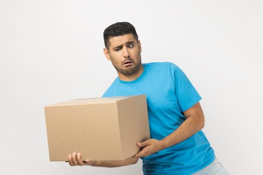 Portrait of funny positive optimistic unshaven man wearing blue T- shirt standing holding very heavy cardboard parcel, making effort to carry box. Indoor studio shot isolated on gray background.