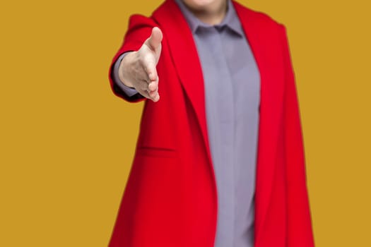 Closeup portrait of unknown woman standing with outstretched hand, saying hello or bye, handshaking, congratulating, wearing red jacket. Indoor studio shot isolated on yellow background.