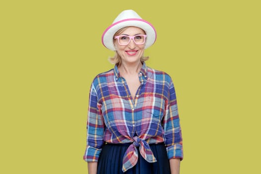 Portrait of smiling joyful senior woman wearing checkered shirt, hat and eyeglasses, looking at camera with toothy smile, expressing positive emotions. Indoor studio shot isolated on yellow background