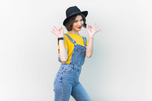 Portrait of optimistic happy hipster woman in blue denim overalls, yellow T-shirt and black hat, raised her arms, showing okay sign. Indoor studio shot isolated on gray background.