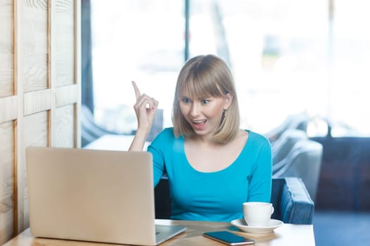 Smart excited positive young woman with blonde hair in blue shirt working on laptop, raised her finger, having idea, knows how to solve problem. Indoor shot in cafe with big window on background.