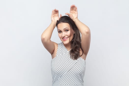 Portrait of positive brunette woman making funny bunny ears with hands on head, childish behavior, optimistic mood, wearing striped dress. Indoor studio shot isolated on gray background.