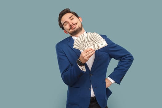 Portrait of rich proud satisfied man with mustache standing showing fan of dollars banknotes, keeps hand on hip, wearing official style suit. Indoor studio shot isolated on light blue background.