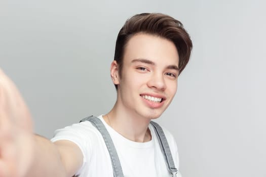 Portrait of optimistic young brunette man standing keeps arms outstretched has optimistic cheerful expression, wearing denim overalls. Indoor studio shot isolated on gray background.