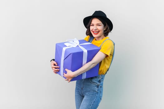 Portrait of extremely happy optimistic hipster woman in blue denim overalls, yellow T-shirt and black hat, embracing big wrapped box with present. Indoor studio shot isolated on gray background.