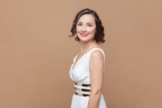 Side view portrait of smiling happy optimistic middle aged woman with wavy hair looking at camera with charming smile, wearing white dress. Indoor studio shot isolated on light brown background.