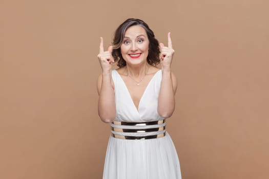 Portrait of smiling happy satisfied middle aged woman with wavy hair pointing upwards and advertisement area, wearing white dress. Indoor studio shot isolated on light brown background.