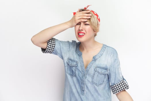 Portrait of sorrowful depressed blonde young woman wearing blue denim shirt and red headband standing making showing facepalm gesture. Indoor studio shot isolated on gray background.
