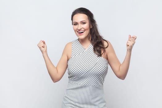 Emotional brunette woman raises clenched fists, exclaims with excitement, rejoices sweet success, feels taste of victory, wearing striped dress. Indoor studio shot isolated on gray background.