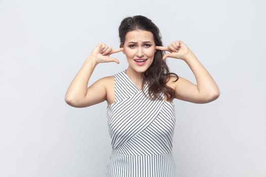 Portrait of brunette woman covering ears as hears very loud noise or music, clenches teeth, ignores something noisy, wearing striped dress. Indoor studio shot isolated on gray background.