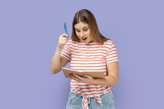 Portrait of excited amazed optimistic blond woman wearing striped T-shirt writing in paper notebook, raised pen, having excellent idea. Indoor studio shot isolated on purple background.
