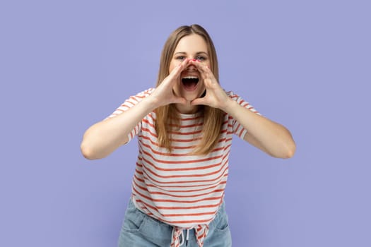 Angry attractive young adult blond woman wearing striped T-shirt standing and screaming with hands near mouth, expressing aggressive emotions. Indoor studio shot isolated on purple background.
