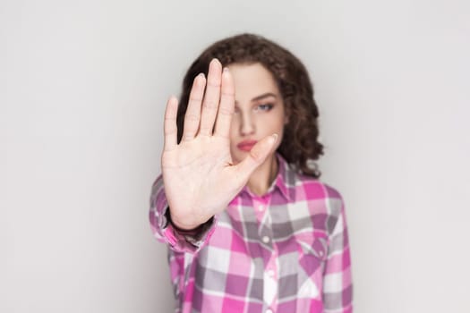Woman with curly hair showing stop sign, prohibition symbol, keeps palm forward to camera, looks with strict expression, wearing pink checkered shirt. Indoor studio shot isolated on gray background.