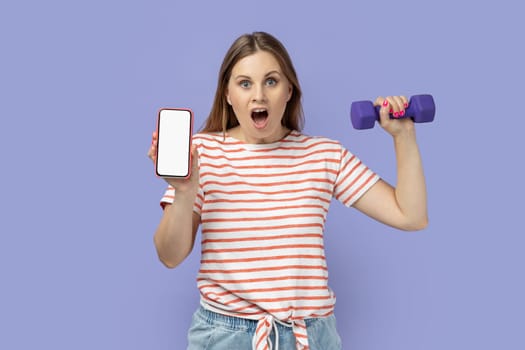 Portrait of amazed surprised blond woman wearing striped T-shirt holding dumbbell in hand and mobile phone with blank screen, space for advertisement. Indoor studio shot isolated on purple background.